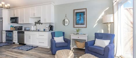 Cocina moderna con armarios blancos, microondas y cafetera. Un área de estar con vistas al exterior, con dos sillones azules y dos otomanas tejidas.