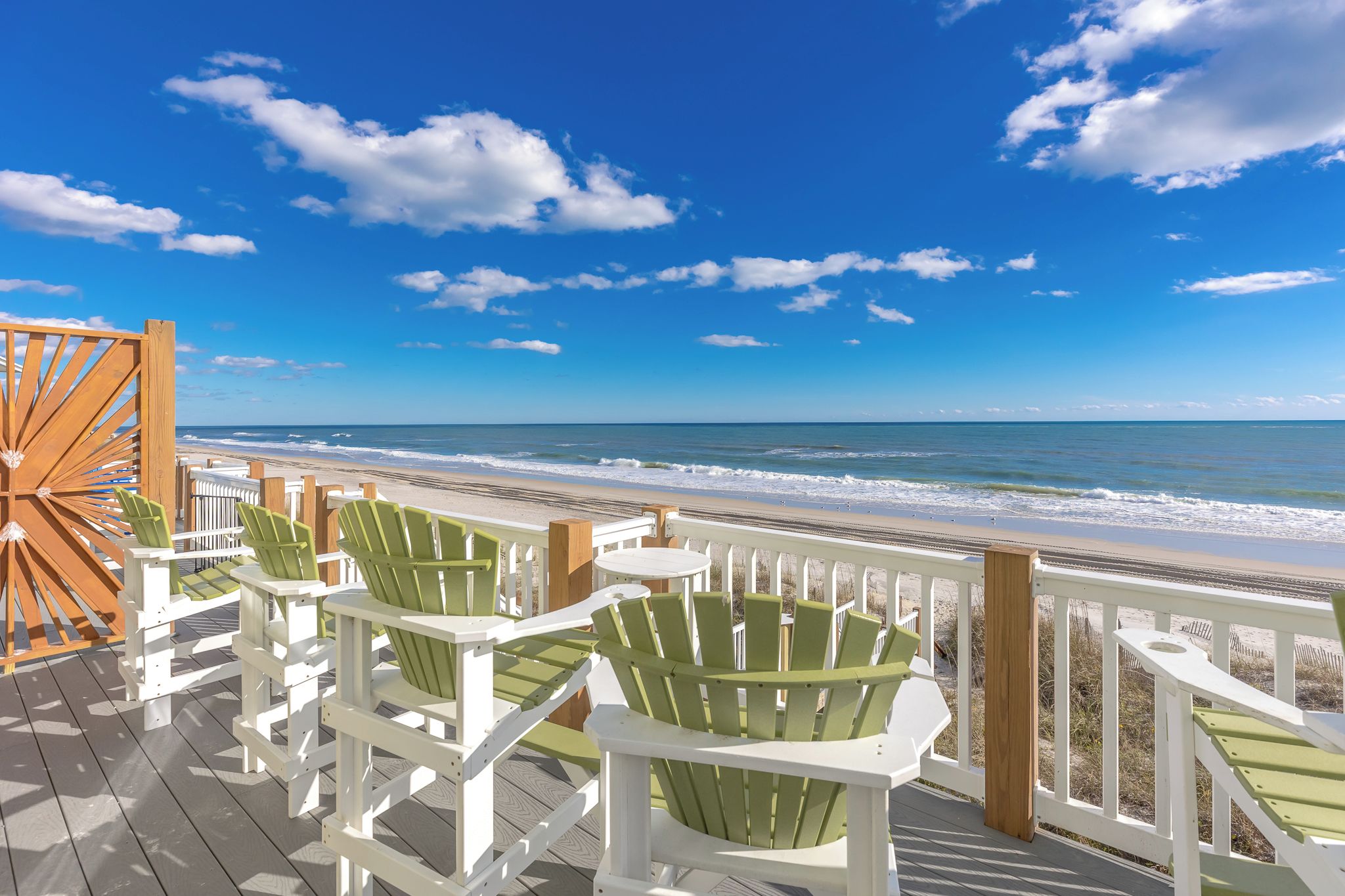 A wooden deck with white and green Adirondack chairs overlooking a beach.