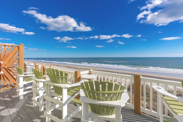 A wooden deck with white and green Adirondack chairs overlooking a beach.