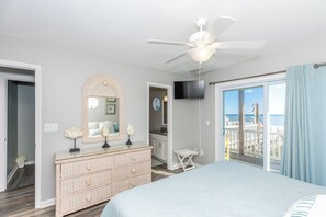 A bedroom with a bed, dresser, mirror, ceiling fan, and a view of the ocean through a balcony.