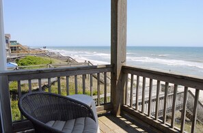 Een houten terras met een rieten stoel en een tafel, met uitzicht op een strand met houten looppaden en een zeezicht.