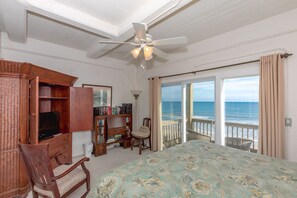 A bedroom with a bed, a wooden wardrobe, a desk, a chair, a bookshelf, and a view of the ocean through a sliding glass door.