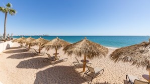 A beach with rows of sun loungers and thatched umbrellas, a palm tree, and a clear view of the ocean. - Sonoran Sea W-109 Serenity By The Sea Bright Oceanfront Condo (Puerto Peñasco)