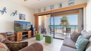 Un soggiorno con TV, mobile TV in legno, pouf verdi e un balcone con vista sulla spiaggia e sull'oceano.