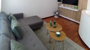 Una sala de estar moderna con un sofá seccional gris, una mesa de centro redonda, un televisor de pantalla plana montado en una pared de madera y un estante blanco pequeño con un ventilador.
