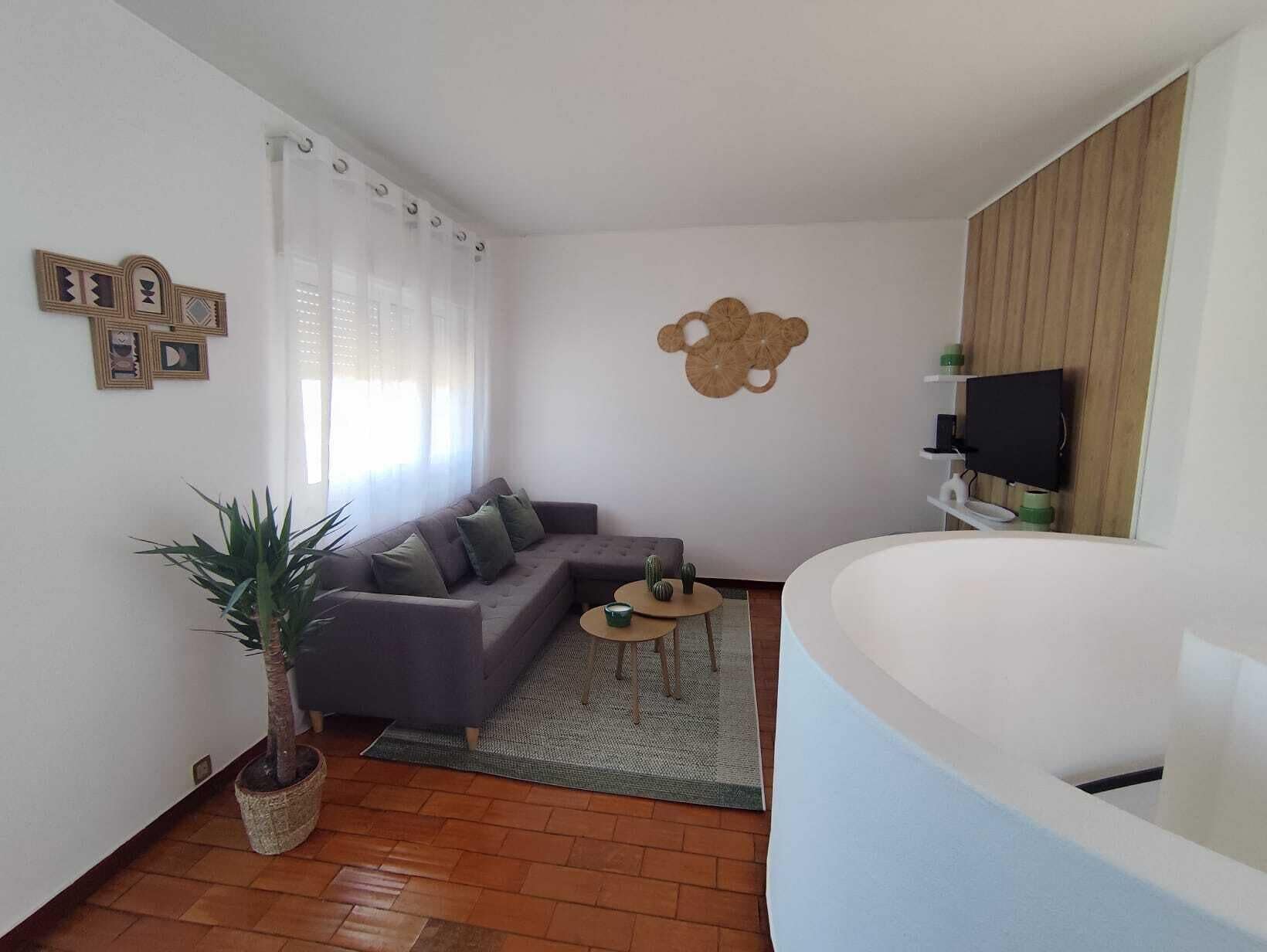 A living room with a sofa, a small round table, a potted plant, and a wall-mounted TV.