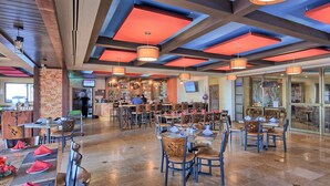 Un restaurant spacieux avec de hauts plafonds, des poutres apparentes et un éclairage moderne suspendu. Les tables sont disposées avec soin et accompagnées de chaises, et un bar est visible au fond de la salle.