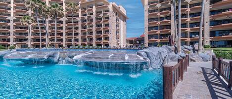 A resort with a large pool featuring multiple water jets, surrounded by palm trees and a multi-story building with balconies.