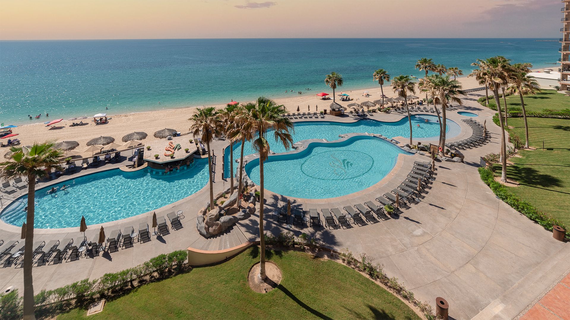 Ein Resort mit großzügiger Poolanlage, Palmen und einem Strand.