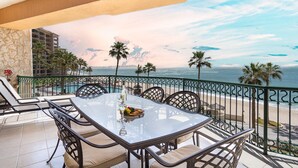 Un balcone con un tavolo e sedie in vetro, vista su una spiaggia con palme e l'oceano.