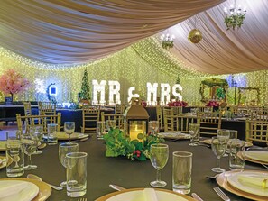 A wedding reception with tables set for guests, featuring a backdrop with 'MR & MRS' letters and festive lighting.