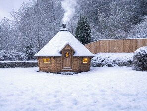 A wooden cabin with a snow-covered roof and a chimney emitting smoke, surrounded by a snowy landscape and a wooden fence. - 16 bedroom accommodation in Bampton, near Tiverton (Bampton, near Tiverton)