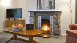 A living room with a stone fireplace, a television on a wooden stand, a bowl of fruit on a table, and a red armchair. - 2 bedroom accommodation in Lostwithiel (Lostwithiel)