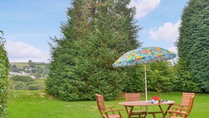 A round wooden table with four chairs, a colorful umbrella, and a view of a green landscape with trees and houses in the background. - 2 bedroom accommodation in Lostwithiel (Lostwithiel)