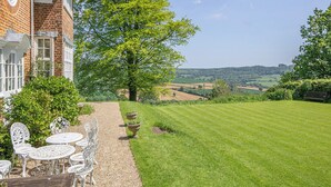 Een bakstenen huis met een tuin, terras en uitzicht op glooiende heuvels.