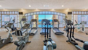 A gym with treadmills, elliptical machines, and weight machines, featuring large windows with a view of the sea.