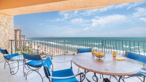 A balcony with a round table, blue chairs, and a view of the beach and ocean.