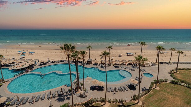 Un resort con una amplia zona de piscina, palmeras y vistas a la playa.