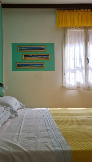 Ein Zimmer mit einem Bett, einem Fenster mit gelben Vorhängen und drei gerahmten Landschaftsbildern an der Wand.