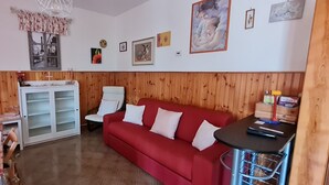 A cozy living room with a red sofa, a wooden panel wall, a white cabinet, and a small table with a chair. - Renovated and equipped apartment in Cervia-Milano Marittima (Cervia)