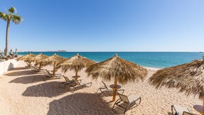 A beach with rows of sun loungers and thatched umbrellas, a palm tree, and a clear view of the ocean.