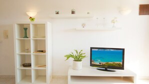 Una sala de estar con un televisor sobre un mueble bajo, una estantería blanca y una planta pequeña.