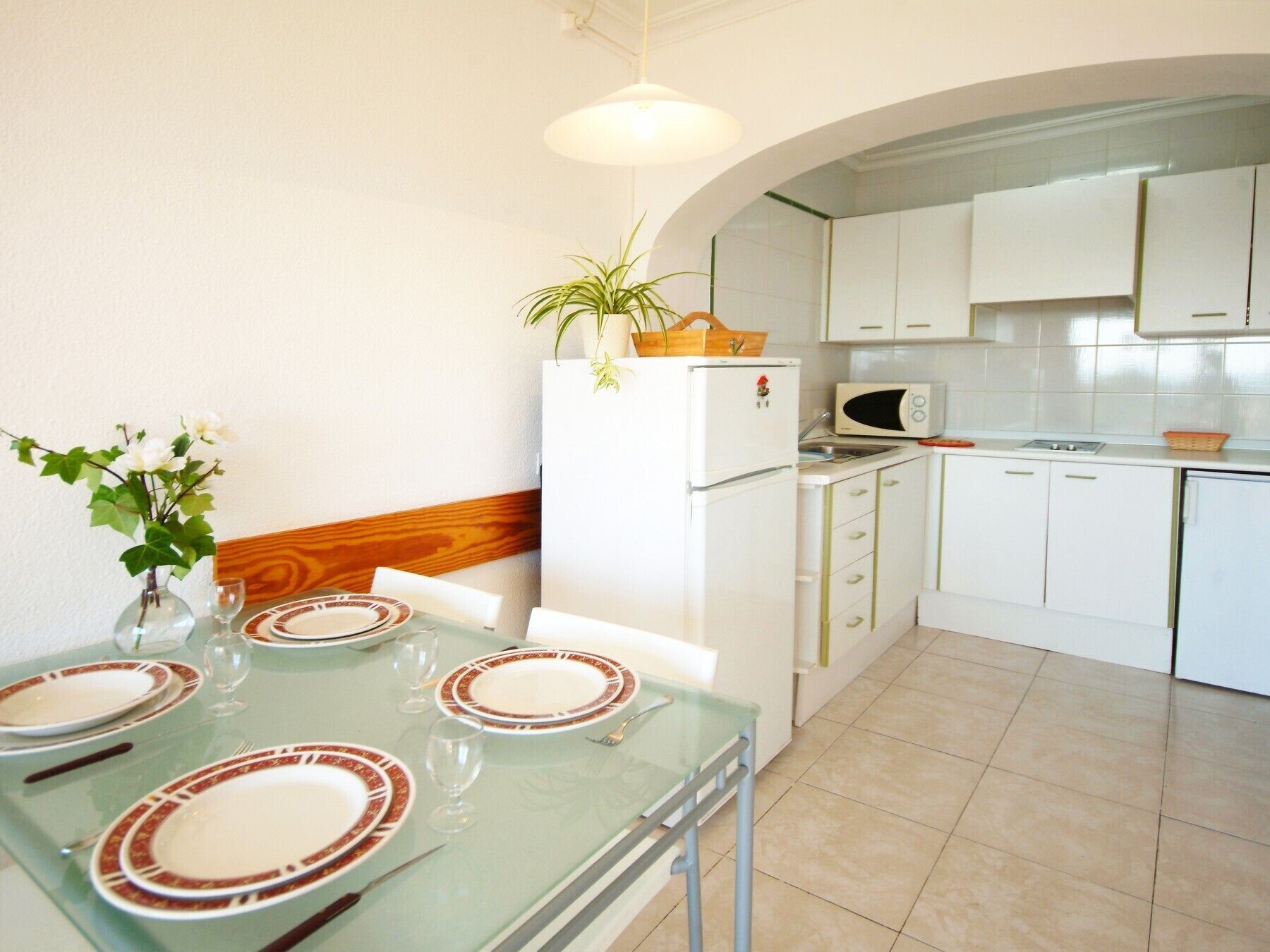 Una cocina compacta con armarios blancos, un microondas y un refrigerador. Hay una mesa de comedor con platos y vasos.