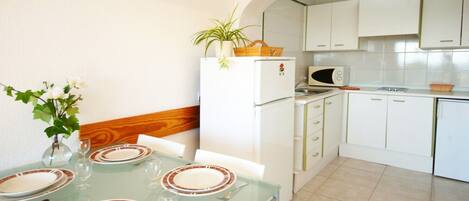 Una cocina compacta con armarios blancos, un microondas y un refrigerador. Hay una mesa de comedor con platos y vasos.