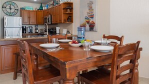 Un coin repas avec une table en bois dressée pour quatre personnes, entourée de chaises en bois. Une grande horloge et une pancarte de bienvenue sont visibles au mur.