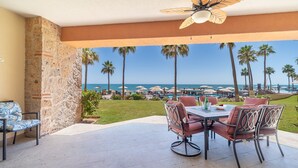 Une terrasse avec une table et des chaises, donnant sur une plage agrémentée de parasols et de palmiers.