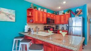 Una cocina con armarios rojos, un refrigerador de acero inoxidable, un microondas y un lavamanos. Hay taburetes de bar y un cuadro en la pared.