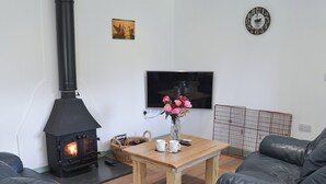 Un salon avec un poêle à bois noir, une table basse en bois sur laquelle est posé un vase de fleurs, un téléviseur fixé au mur et un canapé en cuir noir.
