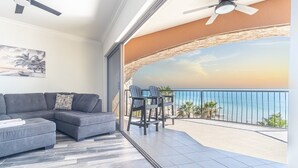 A modern living room with a gray sectional sofa, a small table, and chairs. A large window offers a view of the ocean and palm trees.