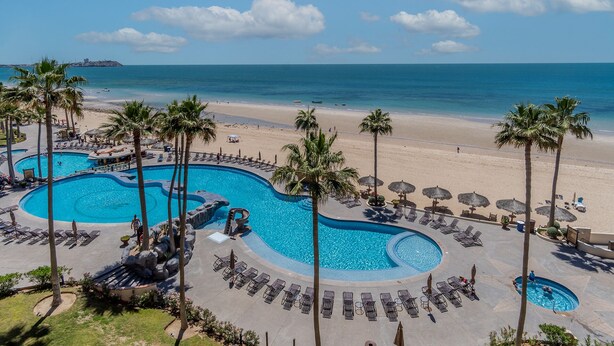 A resort with a large pool area, palm trees, and a beach view.
