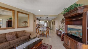 A living room with a sofa, a coffee table, a dining area with chairs, a kitchen in the background, and a TV on a wooden cabinet.