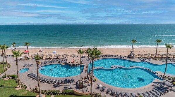 A resort with two large swimming pools, a beach, and palm trees.