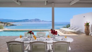 Mesa de comedor lista para cuatro personas, con sillas blancas, vista al mar y una piscina.