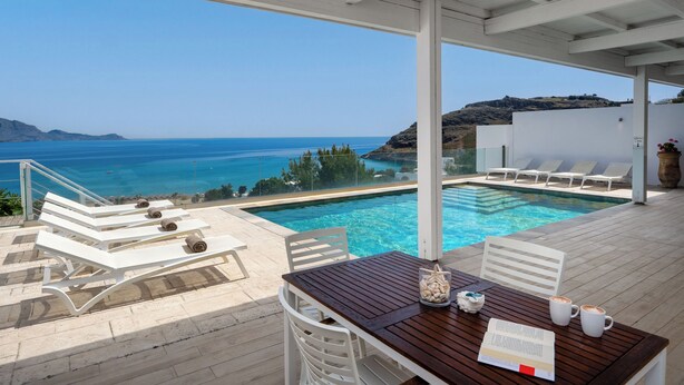 Un área junto a la piscina con sillones de descanso, una mesa de madera y vista al mar.