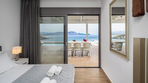 Habitación de hotel con cama, mesita de noche, lámpara, espejo grande y puerta corredera que da a un balcón con piscina y mobiliario exterior.