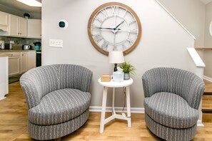A room with two upholstered chairs, a round wall clock, a small white table with a lamp, and a potted plant.