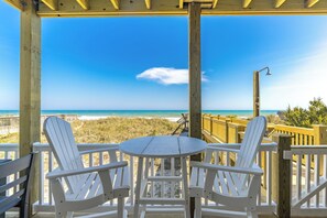 A wooden deck with two white Adirondack chairs and a small round table, overlooking a beach with clear skies. - Misty Morning North - 4BR Oceanfront Home; Dog Friendly! (North Topsail Beach)