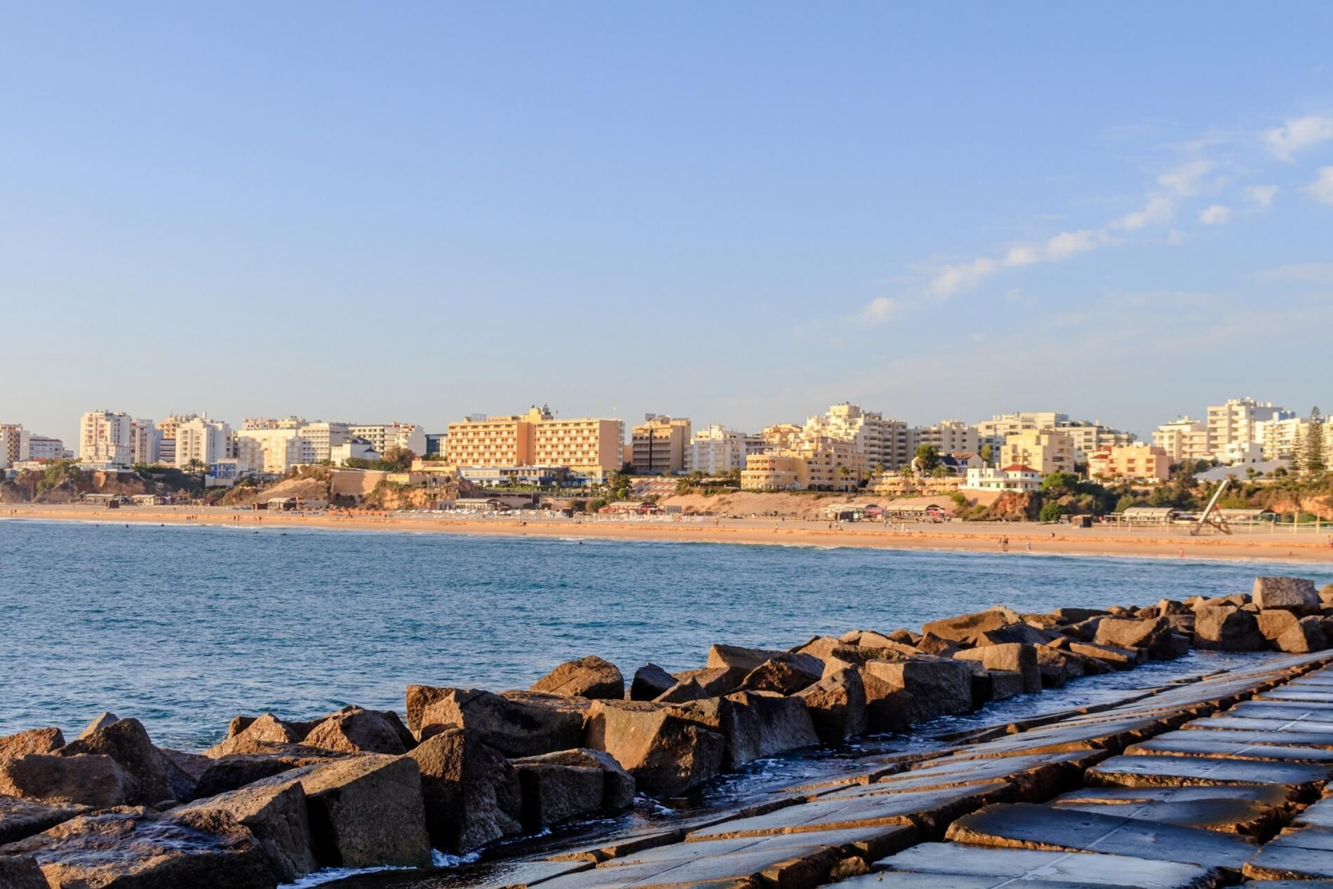 A coastal cityscape with a beach, rocky breakwater, and a row of multi-story buildings.