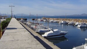 Un port de plaisance avec des bateaux amarrés le long d’une promenade en béton, une digue rocheuse et des montagnes en arrière-plan.
