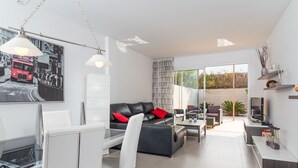 Un salon moderne avec un canapé en cuir noir, des fauteuils blancs, un coin repas avec une table et des chaises blanches, et une grande fenêtre offrant une vue sur l’extérieur.