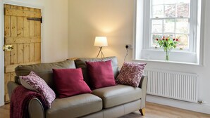 A beige leather sofa with three pillows, a floor lamp, a wooden door, and a window with a view of trees and flowers. - 2 bedroom accommodation in Greenwell, near Brampton (Greenwell, near Brampton)