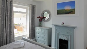 A bedroom with a bed, a dresser, a mirror, a fireplace, and a window with curtains. - Repton Cottage -  a cottage that sleeps 5 guests  in 2 bedrooms (Cromer)