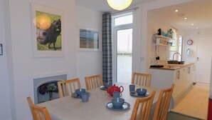 A dining area with a table set for six, surrounded by wooden chairs. There is a fireplace with a decorative mantle, and a window with curtains. - Repton Cottage -  a cottage that sleeps 5 guests  in 2 bedrooms (Cromer)