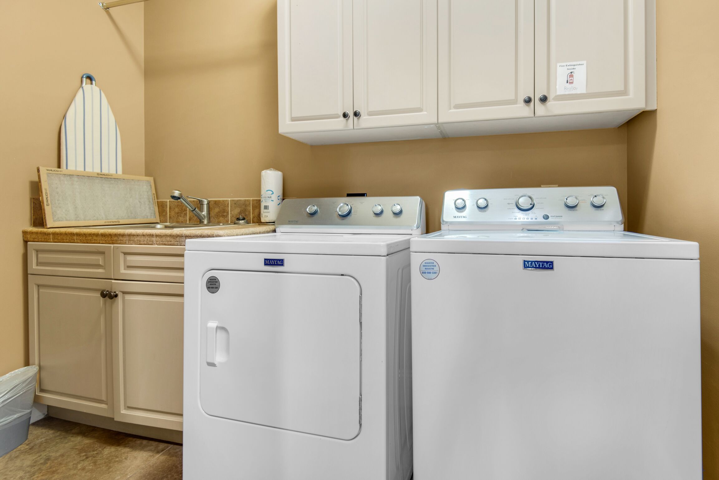 Un lavadero con lavadora y secadora Maytag, armarios color beige y una encimera con dispensador de jabón.
