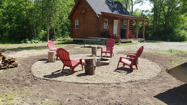 Un chalet rustique doté d’un porche, entouré d’une zone recouverte de gravier, avec des chaises Adirondack rouges et un foyer.