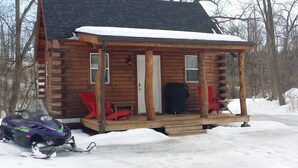 Une cabane en rondins avec un porche, un toit recouvert de neige et une motoneige garée à l’extérieur.